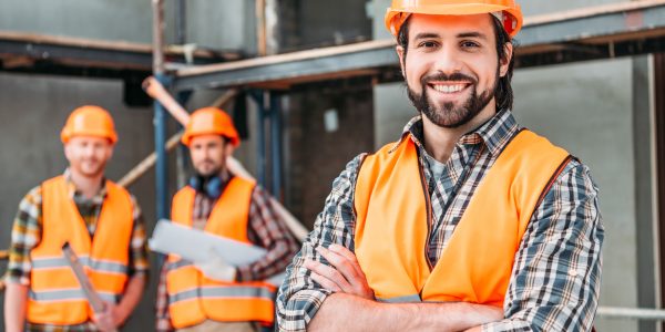 handsome smiling builder standing at construction site with crossed arms while his colleagues